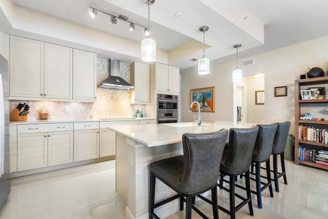 kitchen featuring wall chimney range hood, light tile patterned floors, black electric stovetop, a kitchen island with sink, and backsplash