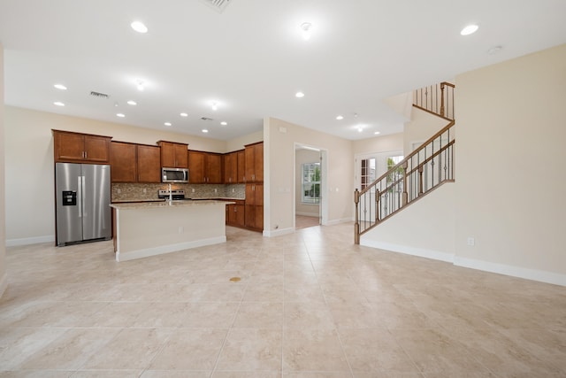 kitchen with a kitchen island with sink, appliances with stainless steel finishes, backsplash, light tile flooring, and light stone countertops