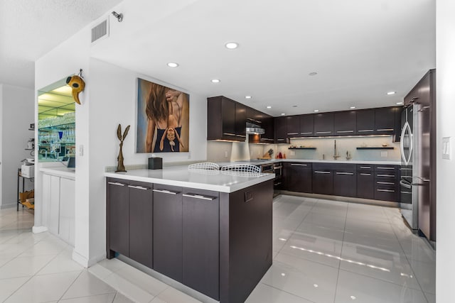 kitchen with backsplash, kitchen peninsula, dark brown cabinets, light tile floors, and stainless steel fridge