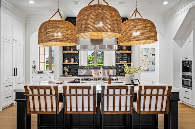 kitchen featuring a kitchen island with sink, exhaust hood, white cabinets, hanging light fixtures, and decorative backsplash