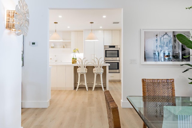 kitchen featuring decorative light fixtures, stainless steel double oven, white cabinetry, light wood-type flooring, and a breakfast bar