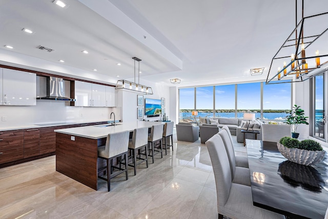 kitchen with a center island with sink, wall chimney range hood, decorative light fixtures, sink, and white cabinetry