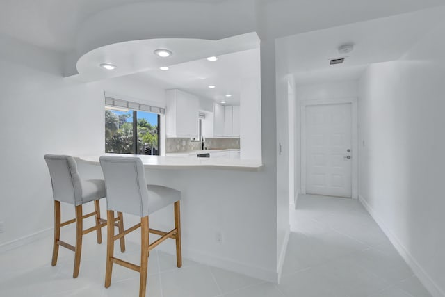 kitchen with kitchen peninsula, a kitchen bar, backsplash, light tile flooring, and white cabinets