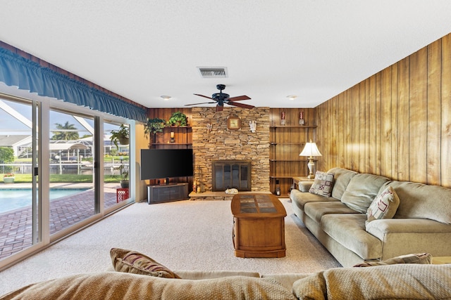 carpeted living room featuring wooden walls, a stone fireplace, and ceiling fan