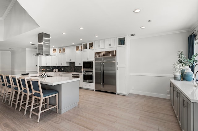 kitchen featuring white cabinets, tasteful backsplash, island range hood, crown molding, and built in appliances