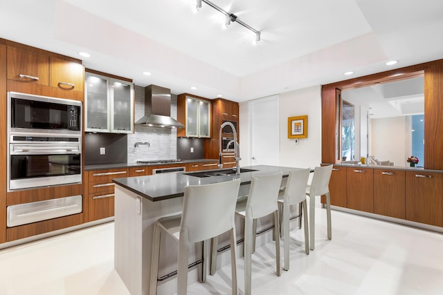 kitchen featuring stainless steel appliances, tasteful backsplash, an island with sink, wall chimney exhaust hood, and a kitchen breakfast bar