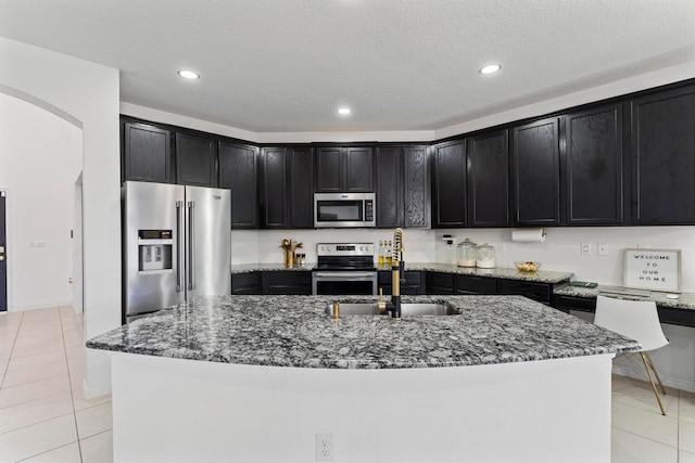 kitchen featuring an island with sink, sink, dark stone counters, light tile patterned floors, and stainless steel appliances