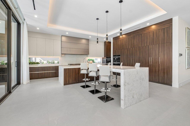 kitchen featuring custom range hood, a raised ceiling, wooden walls, pendant lighting, and a center island with sink