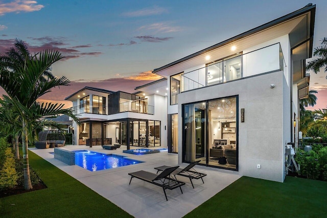 back house at dusk with a swimming pool with hot tub, a patio, a balcony, and a yard