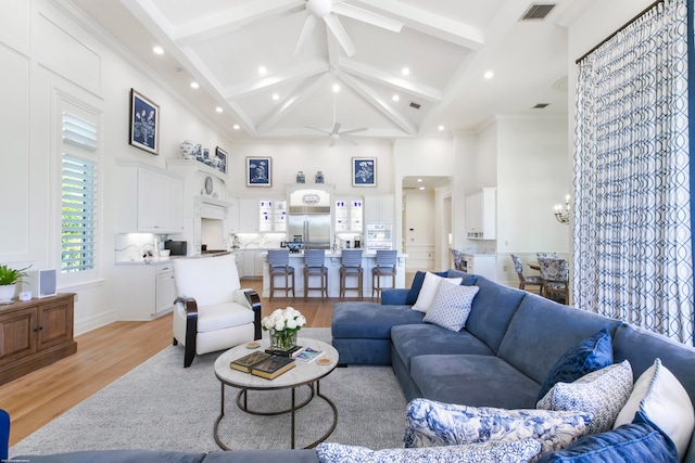 living room with beamed ceiling, ceiling fan, light wood-type flooring, and a high ceiling