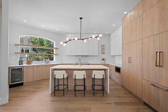 kitchen with decorative light fixtures, light hardwood / wood-style flooring, beverage cooler, a kitchen island with sink, and white cabinets