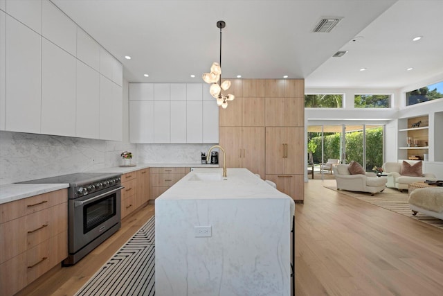 kitchen featuring decorative light fixtures, backsplash, stainless steel range, light hardwood / wood-style floors, and an island with sink