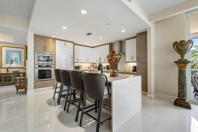 kitchen featuring a kitchen breakfast bar, an island with sink, light tile flooring, white cabinetry, and appliances with stainless steel finishes