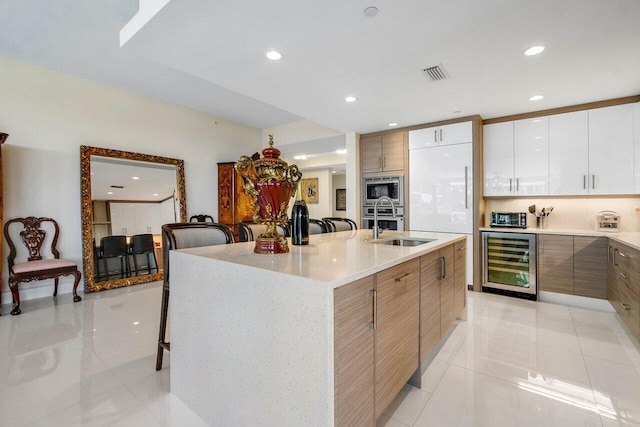 kitchen with stainless steel microwave, white cabinets, a kitchen island with sink, wine cooler, and light tile floors