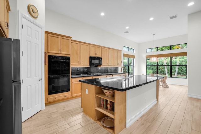 kitchen with a center island with sink, light wood-type flooring, black appliances, dark stone counters, and sink