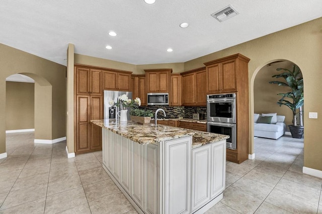 kitchen with appliances with stainless steel finishes, backsplash, light stone counters, a kitchen island with sink, and light tile patterned floors