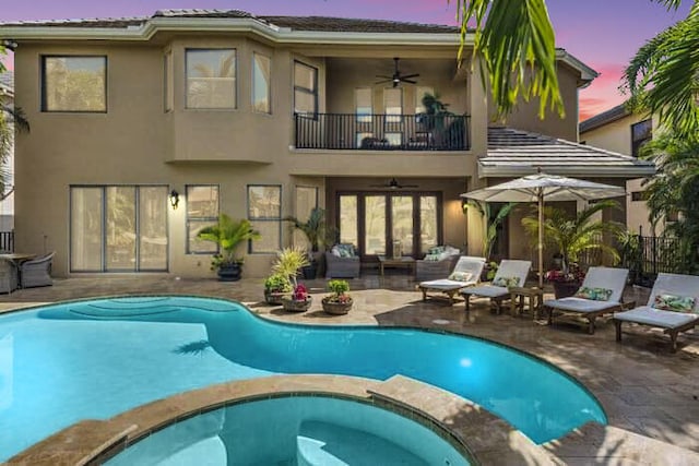 back house at dusk featuring a balcony, ceiling fan, a patio area, and a pool with hot tub