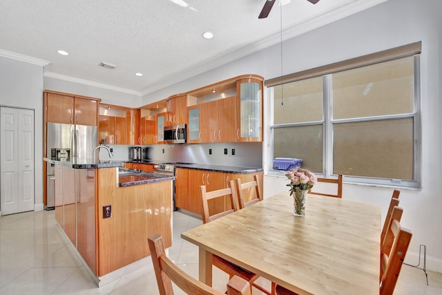 kitchen with ceiling fan, appliances with stainless steel finishes, hanging light fixtures, ornamental molding, and a kitchen island with sink