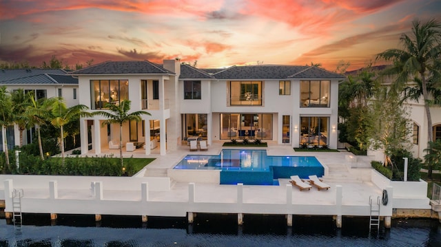 back house at dusk with a patio area, a balcony, and a water view