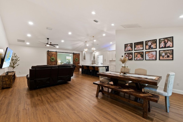 dining space with wood-type flooring, vaulted ceiling, ceiling fan with notable chandelier, and a barn door