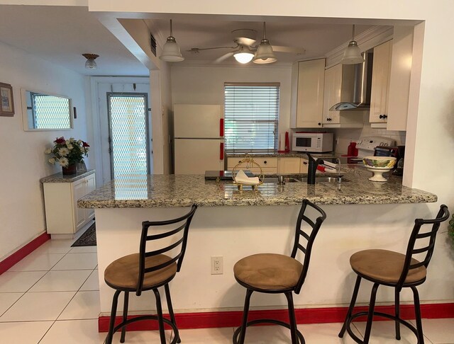 kitchen featuring wall chimney range hood, kitchen peninsula, a wealth of natural light, white appliances, and light tile floors