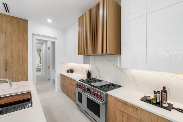 kitchen featuring light tile floors, backsplash, wall chimney range hood, range with two ovens, and light stone countertops