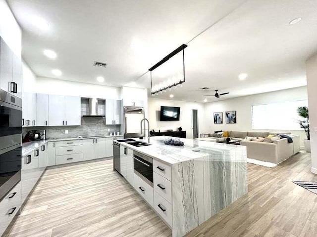 kitchen with light wood-type flooring, sink, an island with sink, white cabinets, and wall chimney range hood