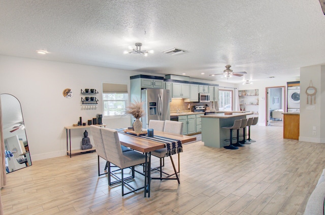 dining space with ceiling fan with notable chandelier, a textured ceiling, light wood-type flooring, and stacked washer and clothes dryer