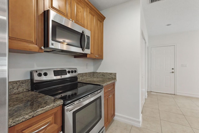 kitchen with light tile patterned floors, brown cabinets, stainless steel appliances, and dark stone counters