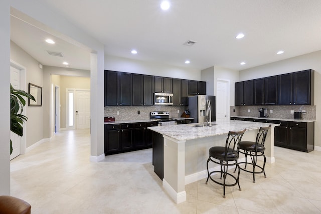 kitchen featuring backsplash, appliances with stainless steel finishes, an island with sink, and light stone counters