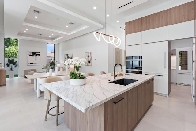 kitchen featuring a tray ceiling, a kitchen island with sink, sink, decorative light fixtures, and white cabinetry