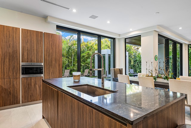 kitchen with dark stone counters, a center island with sink, oven, light tile floors, and sink