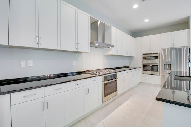 kitchen with stainless steel appliances, light tile flooring, dark stone countertops, wall chimney range hood, and white cabinetry