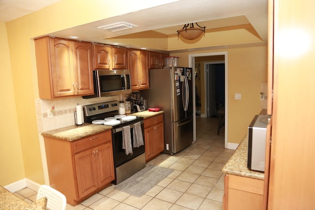 kitchen with a tray ceiling, light stone counters, backsplash, light tile floors, and appliances with stainless steel finishes