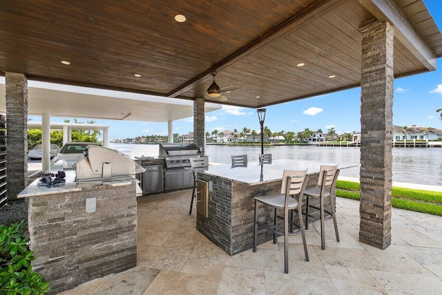 view of patio with a grill, a water view, a bar, ceiling fan, and exterior kitchen