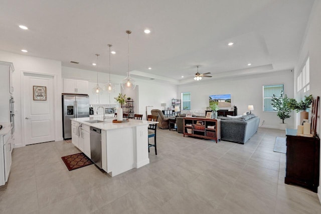 kitchen with ceiling fan, a tray ceiling, an island with sink, sink, and appliances with stainless steel finishes