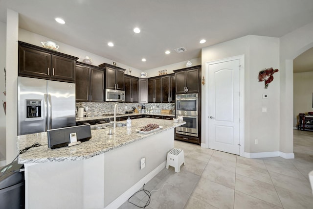 kitchen featuring a center island with sink, appliances with stainless steel finishes, dark brown cabinets, light stone countertops, and decorative backsplash