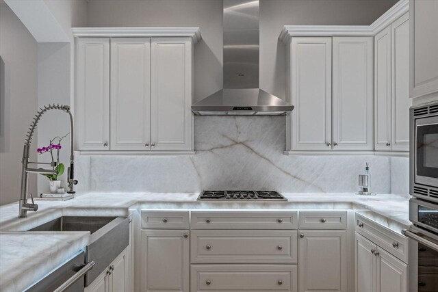 kitchen with wall chimney exhaust hood, backsplash, sink, white cabinetry, and appliances with stainless steel finishes