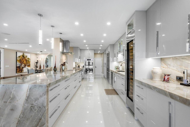 kitchen featuring wall chimney range hood, stainless steel double oven, decorative light fixtures, and light tile floors