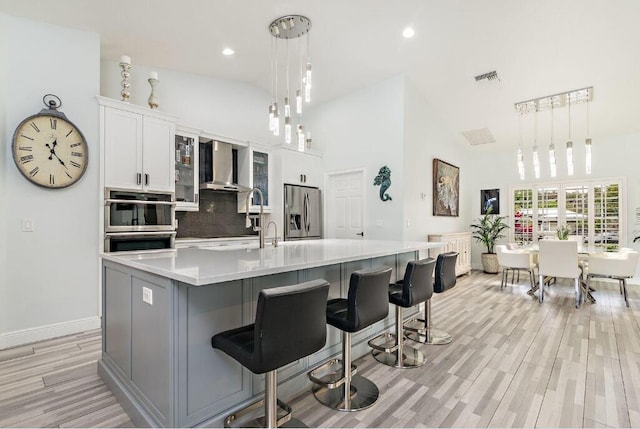 kitchen featuring light hardwood / wood-style floors, appliances with stainless steel finishes, wall chimney exhaust hood, a large island with sink, and high vaulted ceiling