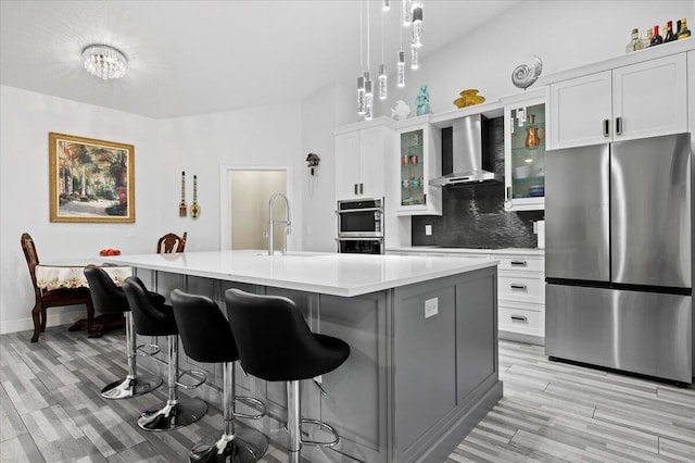 kitchen with hanging light fixtures, a center island with sink, stainless steel appliances, wall chimney exhaust hood, and backsplash