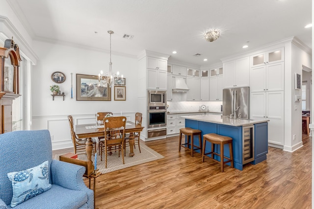 kitchen featuring premium range hood, decorative light fixtures, a center island with sink, white cabinets, and appliances with stainless steel finishes