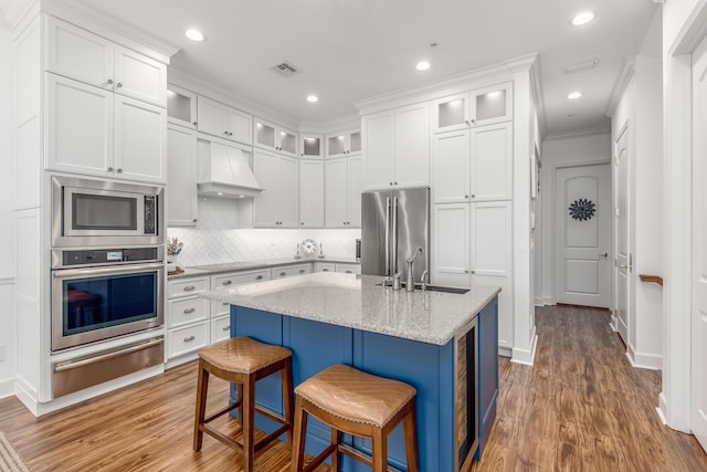 kitchen featuring custom exhaust hood, stainless steel appliances, sink, a center island with sink, and white cabinets