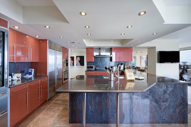 kitchen featuring wall chimney range hood, dark tile floors, backsplash, stainless steel appliances, and a raised ceiling