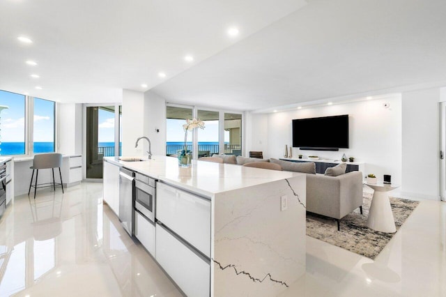 kitchen with a water view, an island with sink, light tile floors, sink, and white cabinets