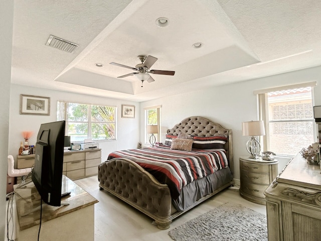 bedroom with ceiling fan, a textured ceiling, a tray ceiling, and light wood-type flooring