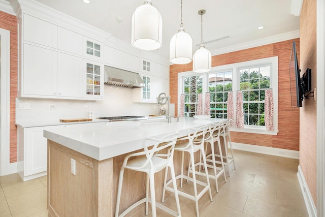 kitchen featuring wall chimney range hood, decorative light fixtures, white cabinets, and an island with sink