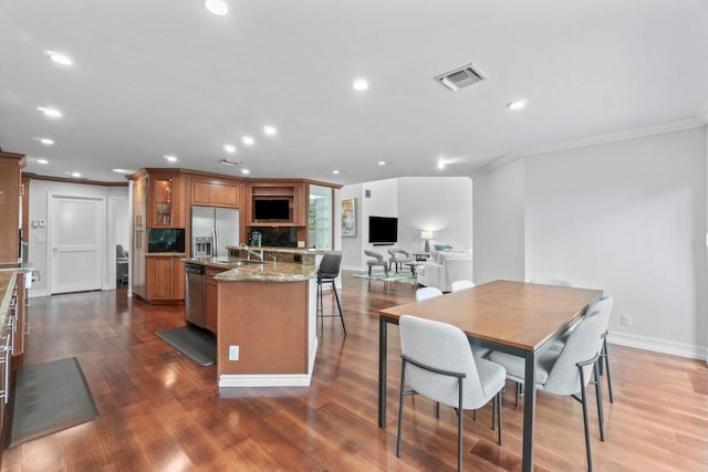 kitchen with a kitchen island with sink, dark wood-type flooring, crown molding, light stone counters, and stainless steel appliances