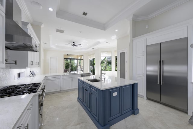 kitchen with a tray ceiling, a kitchen island with sink, wall chimney range hood, high quality appliances, and white cabinets