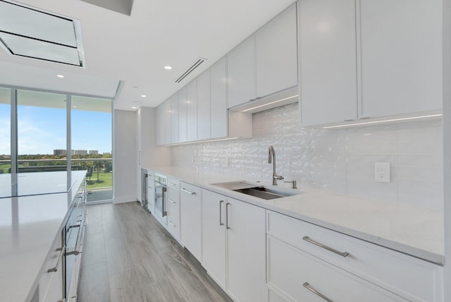 kitchen with sink, light hardwood / wood-style flooring, a wall of windows, backsplash, and white cabinets